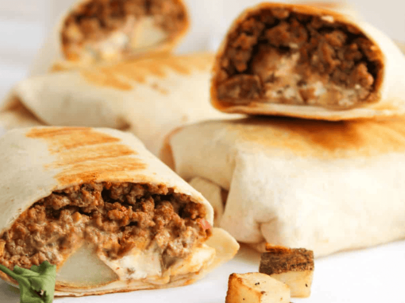 Close-up of several wrapped burritos on a table, with some cut open showing a filling of ground meat and sauce. Pieces of roasted potato and cilantro are scattered around.