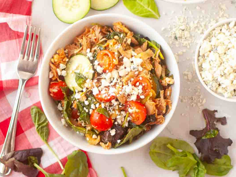 A bowl of salad with shredded chicken, grape tomatoes, cucumber, leafy greens, and crumbled cheese sits on a white surface next to a fork.