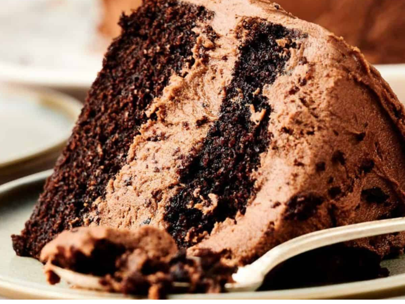 A close-up of a slice of chocolate layer cake with chocolate frosting on a plate, with a fork beside it.
