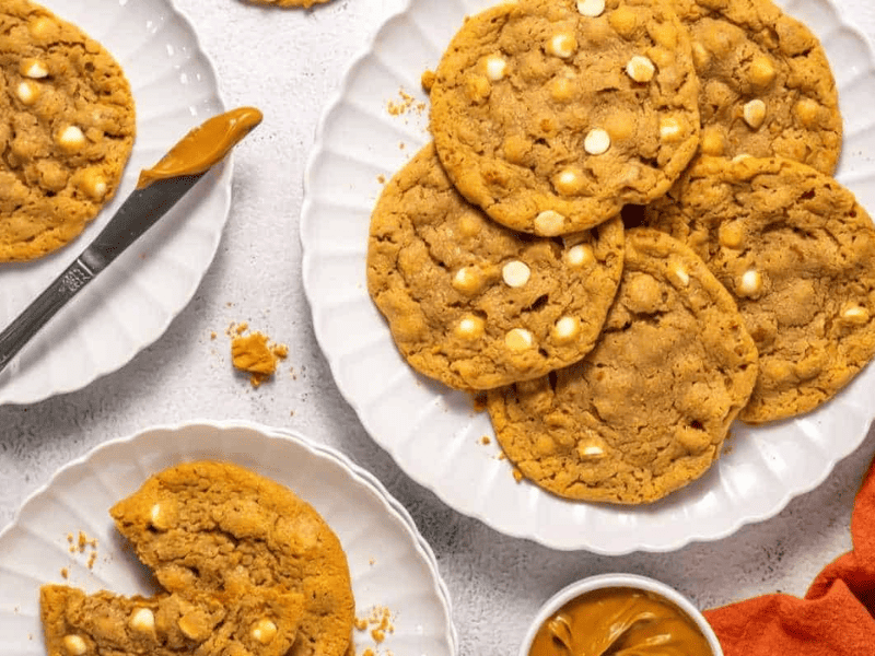 A plate of large peanut Copycat Crumbl Biscoff cookies with white chocolate chips, surrounded by a knife, a small bowl of peanut butter, and an orange napkin.