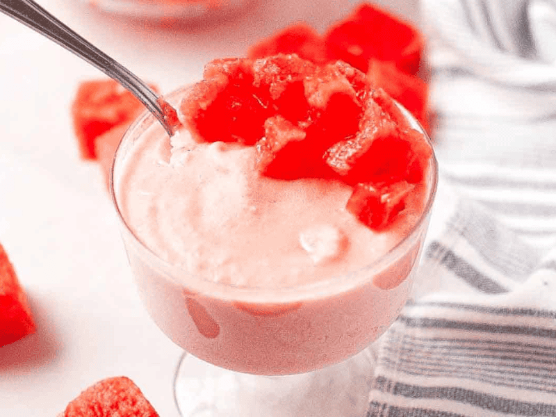A glass dish filled with pink watermelon mousse, topped with diced watermelon pieces, sits on a white surface next to a striped cloth.