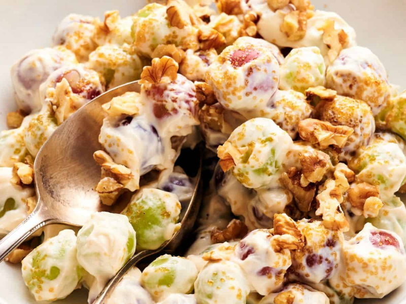 A bowl of grape salad with green and red grapes coated in creamy dressing, topped with chopped walnuts and brown sugar, with a spoon for serving.