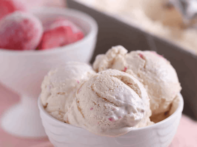 Three scoops of strawberry ice cream in a white bowl, with a bowl of frozen strawberries and an ice cream container in the background.