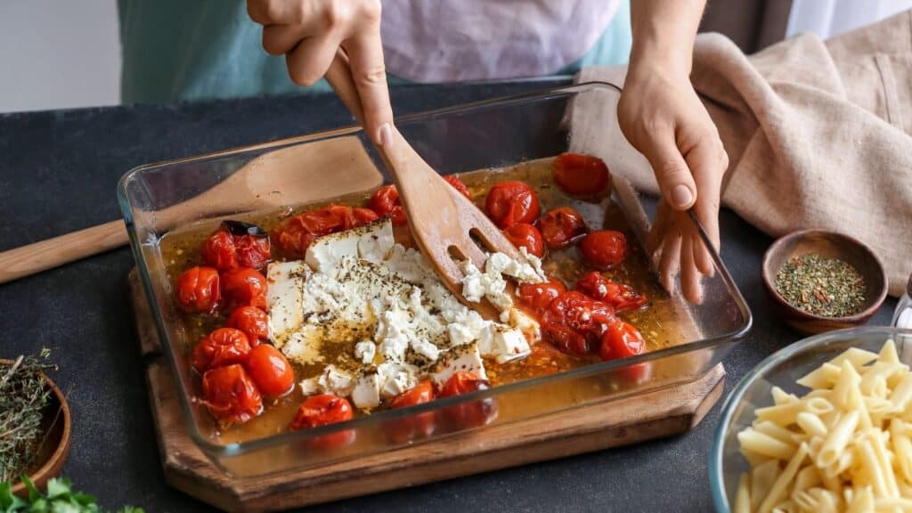 A person uses wooden utensils to mix baked feta cheese and cherry tomatoes in a glass dish, with pasta and seasonings nearby.