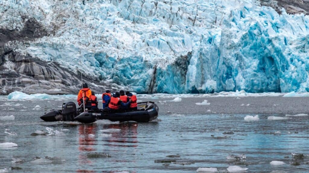 A group of people in orange life vests ride a small inflatable boat near a blue glacier, floating among ice chunks on cold water—a thrilling excursion on a Windstar Alaska cruise.