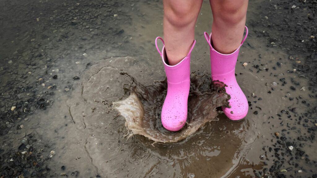 Child in pink rain boots splashing in a muddy puddle on wet pavement, celebrating International Mud Day.