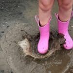 Child in pink rain boots splashing in a muddy puddle on wet pavement, celebrating International Mud Day.