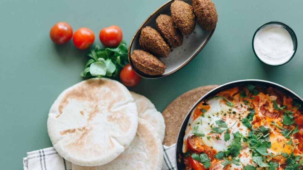 A table set for International Falafel Day features pita bread, fresh tomatoes, greens, a bowl of falafel, a small cup of white sauce, and a skillet of shakshuka garnished with herbs.