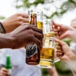 Several people clink beer bottles and mugs together in a happy hour toast outdoors, with blurred greenery in the background.