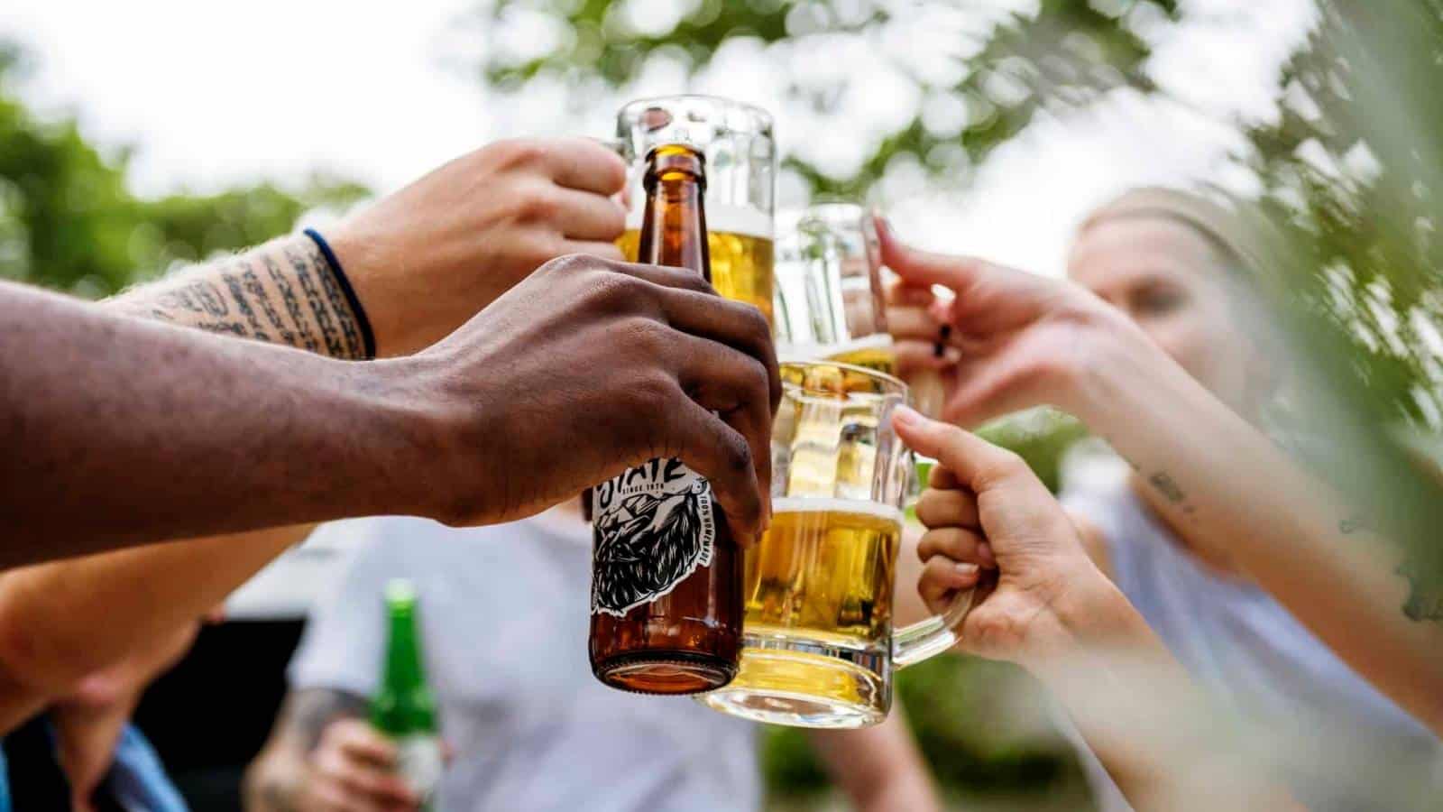 Several people clink beer bottles and mugs together in a happy hour toast outdoors, with blurred greenery in the background.