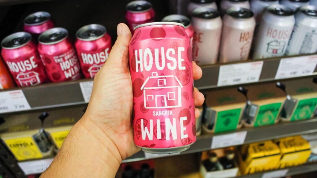 A hand holds a pink can of House Wine Sangria, showcasing the convenience of wine in a can, in front of a store shelf stocked with various canned beverages.
