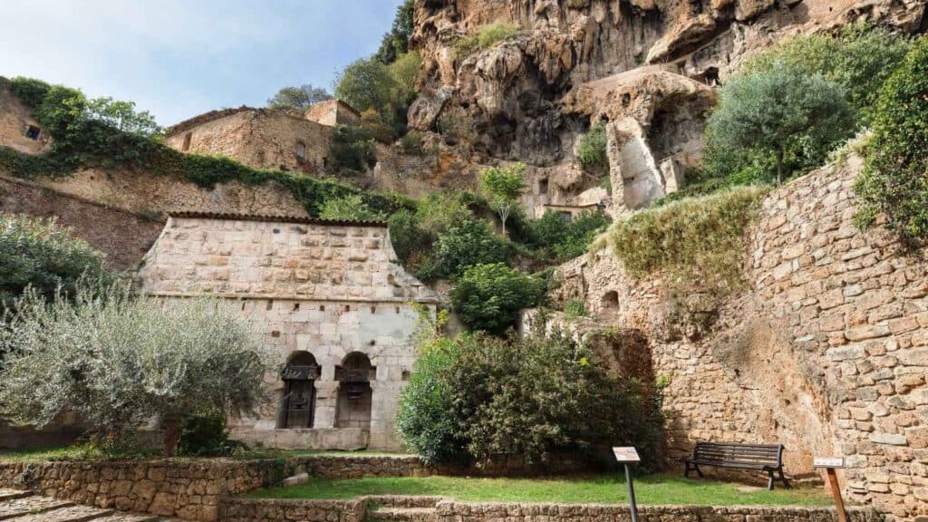 Ancient stone building with arched doorways set against a rocky hillside, surrounded by lush greenery and stone walls.