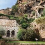 Ancient stone building with arched doorways set against a rocky hillside, surrounded by lush greenery and stone walls.