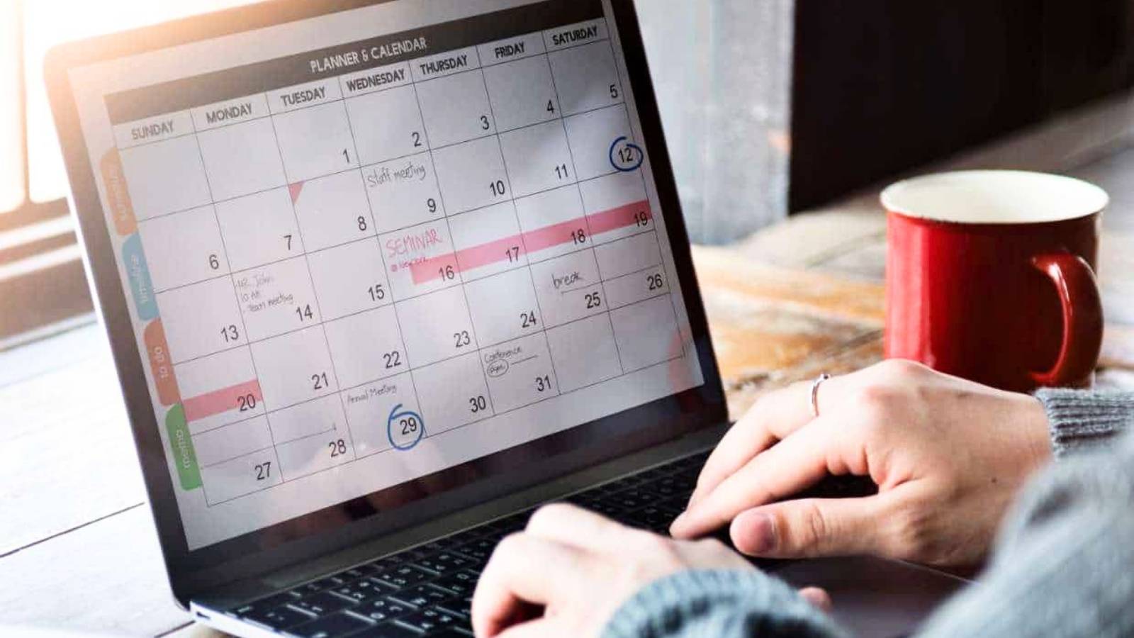 A person types on a laptop displaying a digital monthly calendar, next to a red coffee mug on a wooden table.