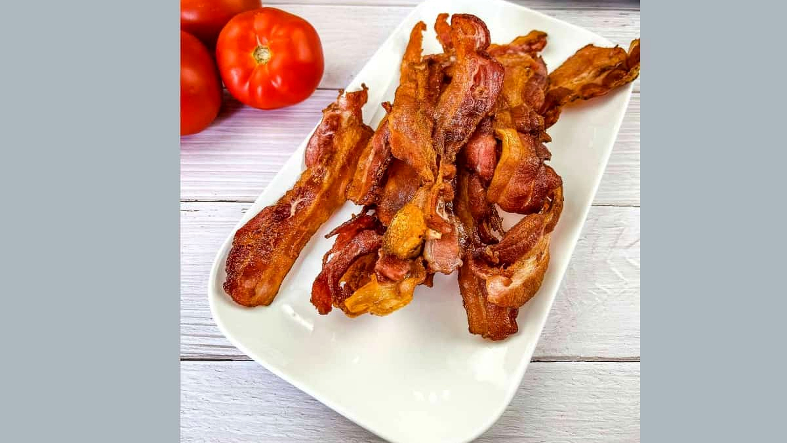 Bacon strips on a white plate next to tomatoes.