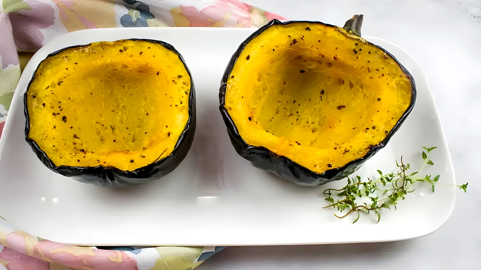 Two roasted acorn squash halves are placed on a white rectangular plate, garnished with a sprig of fresh herbs.