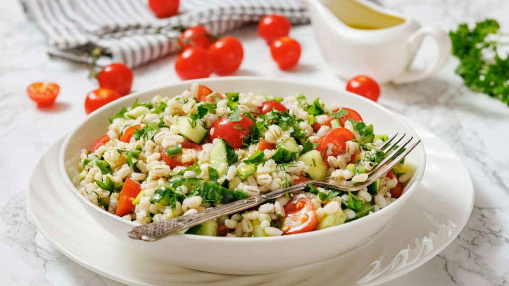A bowl of barley salad with chopped tomatoes, cucumbers, parsley, and a fork, placed on a plate; cherry tomatoes and a sauce pitcher are in the background.