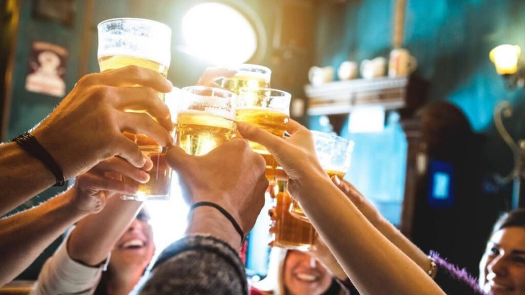 A group of people raise glasses of beer in a toast during happy hour inside a dimly lit bar or pub.
