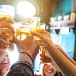 A group of people raise glasses of beer in a toast during happy hour inside a dimly lit bar or pub.