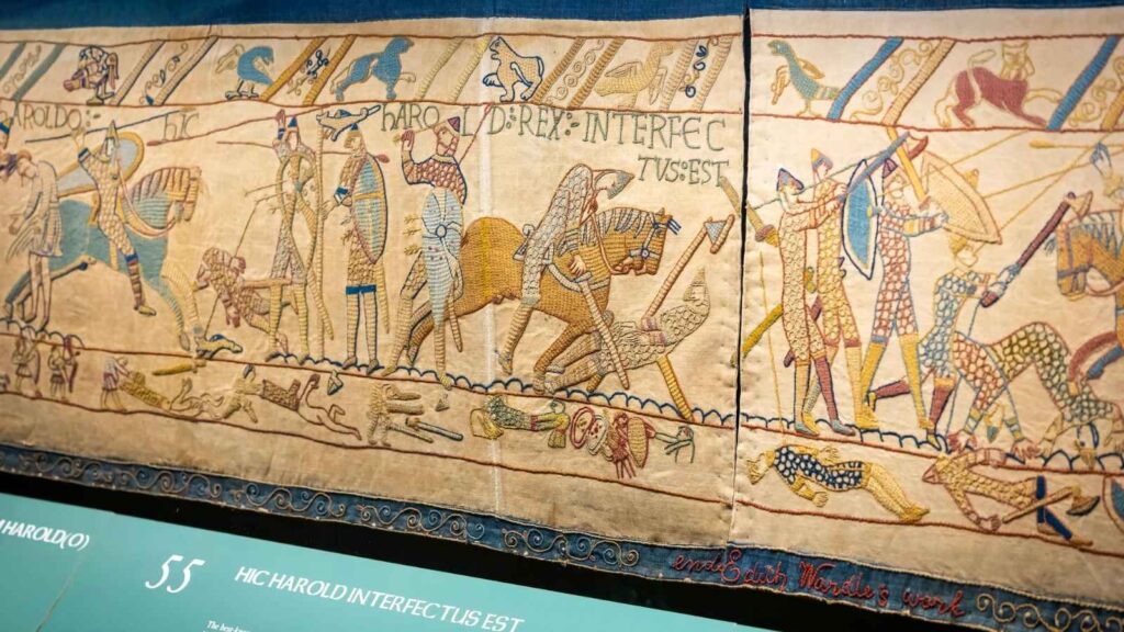 Section of the Bayeux Tapestry depicting a medieval battle scene with soldiers, horses, and weapons, accompanied by Latin inscriptions.
