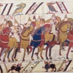A scene from the Bayeux Tapestry showing armored soldiers on horseback, carrying spears and shields, with decorative borders featuring animals and birds.