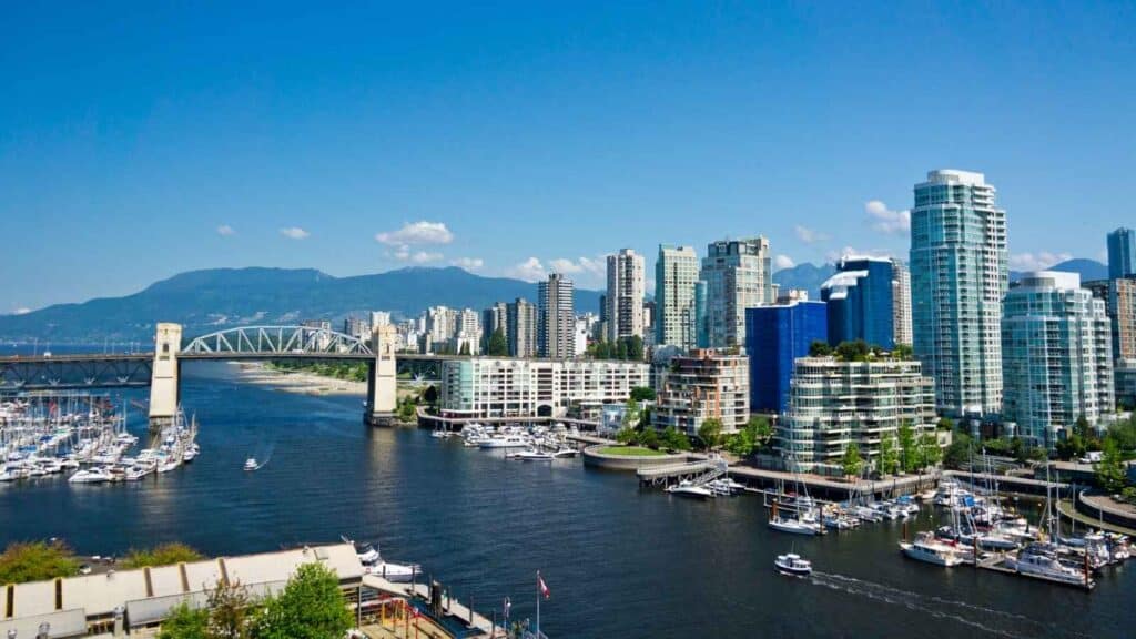 A vibrant cityscape of Vancouver features modern high-rise buildings, a marina with boats, and a bridge spanning the water, with mountains rising in the background beneath a clear blue sky.