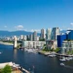 A vibrant cityscape of Vancouver features modern high-rise buildings, a marina with boats, and a bridge spanning the water, with mountains rising in the background beneath a clear blue sky.