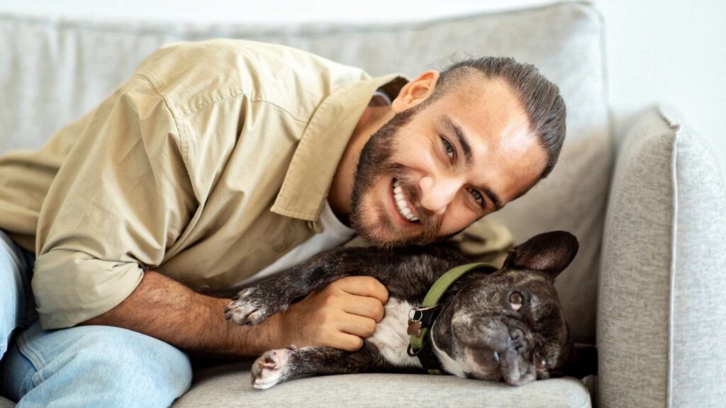 A man with a beard smiles at the camera while lying on a couch next to his black French bulldog in a green collar, capturing a joyful moment perfect for National Dog Dad Day.