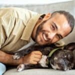 A man with a beard smiles at the camera while lying on a couch next to his black French bulldog in a green collar, capturing a joyful moment perfect for National Dog Dad Day.