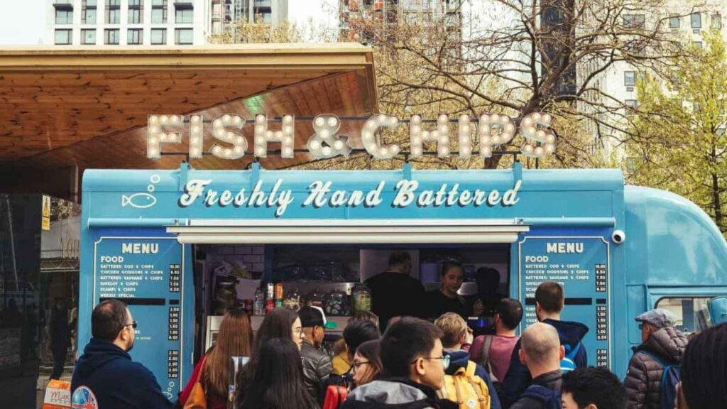 Blue food truck labeled "Fish & Chips Freshly Hand Battered" serves customers; a crowd waits in front, and menus are visible on each side of the counter.