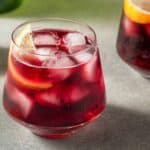 Two glasses of iced red beverage with orange slices sit on a gray surface, next to a halved lemon and a lemon wedge.