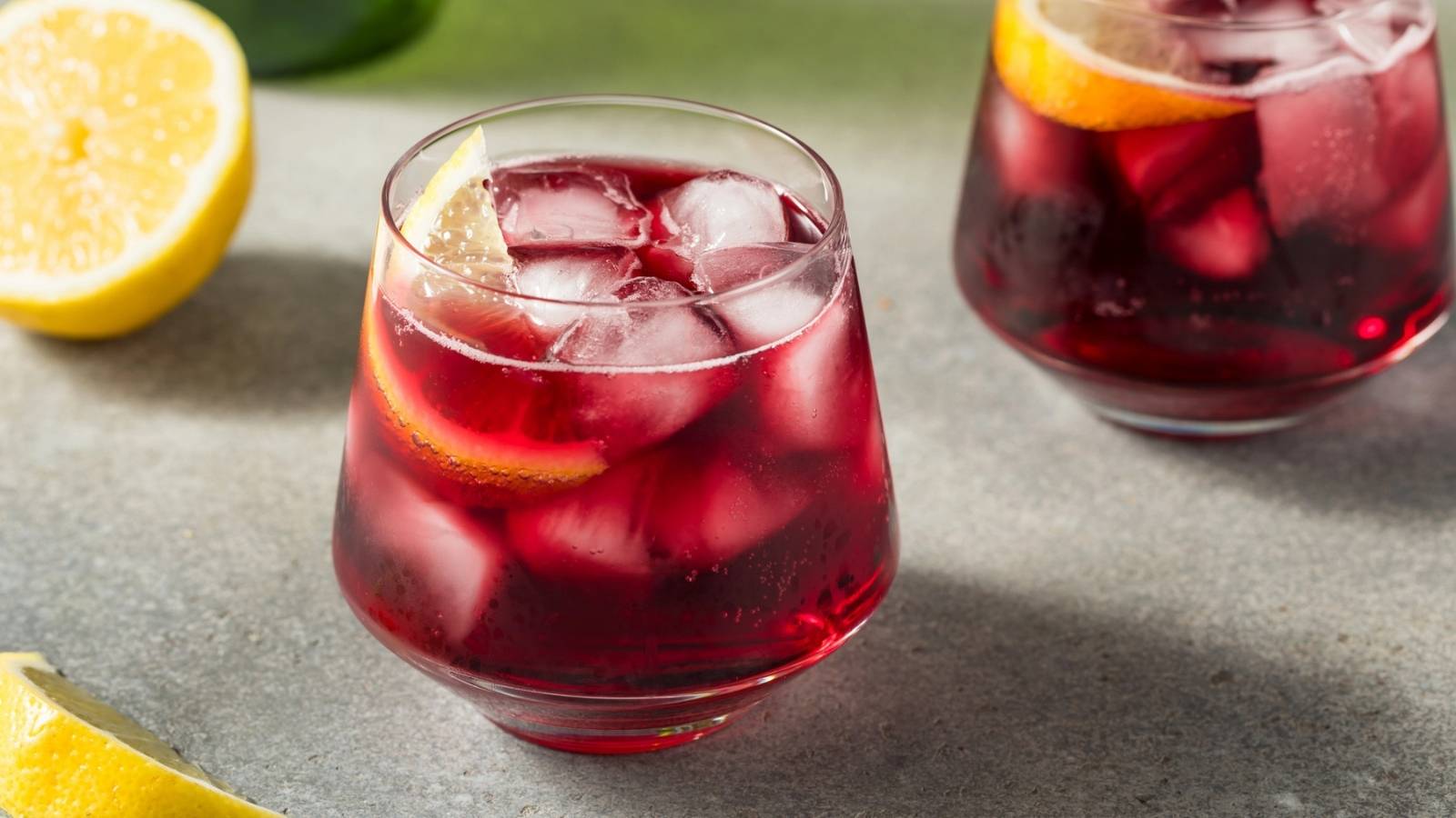 Two glasses of iced red beverage with orange slices sit on a gray surface, next to a halved lemon and a lemon wedge.