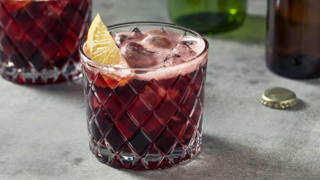 A glass of iced red beverage garnished with a lemon wedge, set on a gray surface with a bottle cap and bottles in the background.