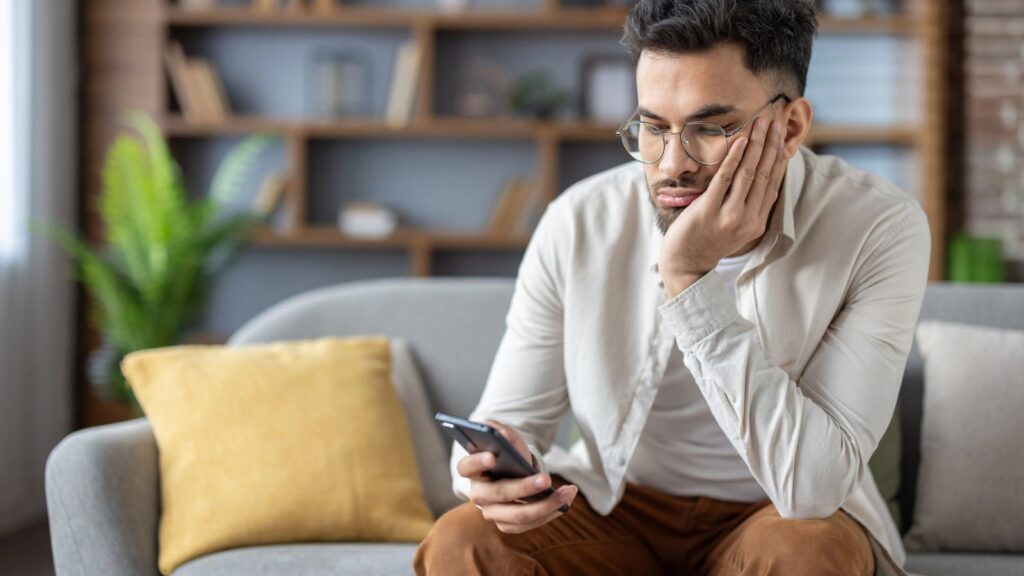A man sits on a couch, resting his face on his hand and looking at his smartphone with a neutral expression&mdash;perhaps reflecting on the friendship recession. A bookshelf and a plant are in the background.