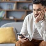 A man sits on a couch, resting his face on his hand and looking at his smartphone with a neutral expression—perhaps reflecting on the friendship recession. A bookshelf and a plant are in the background.