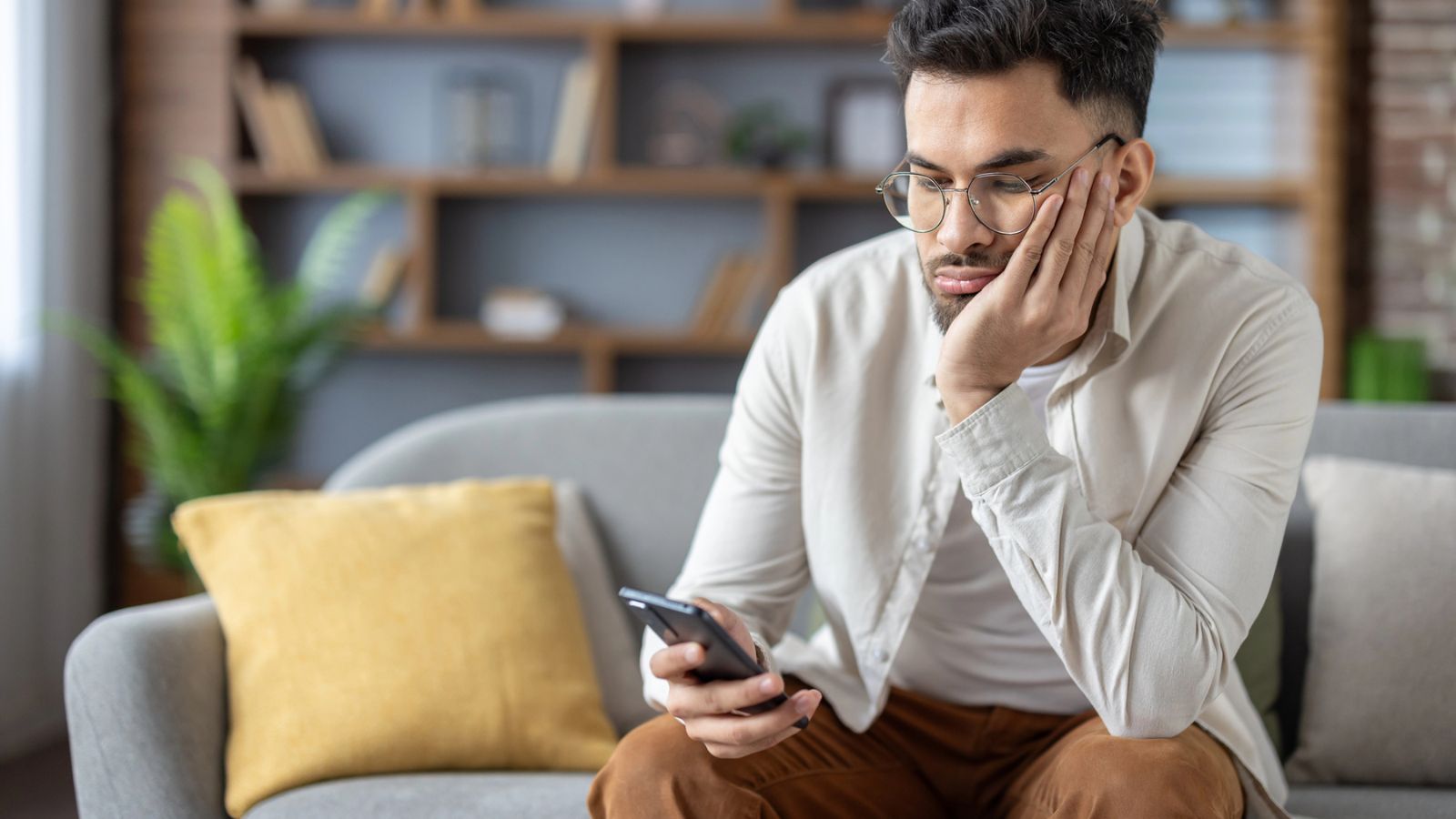A man sits on a couch, resting his face on his hand and looking at his smartphone with a neutral expression—perhaps reflecting on the friendship recession. A bookshelf and a plant are in the background.