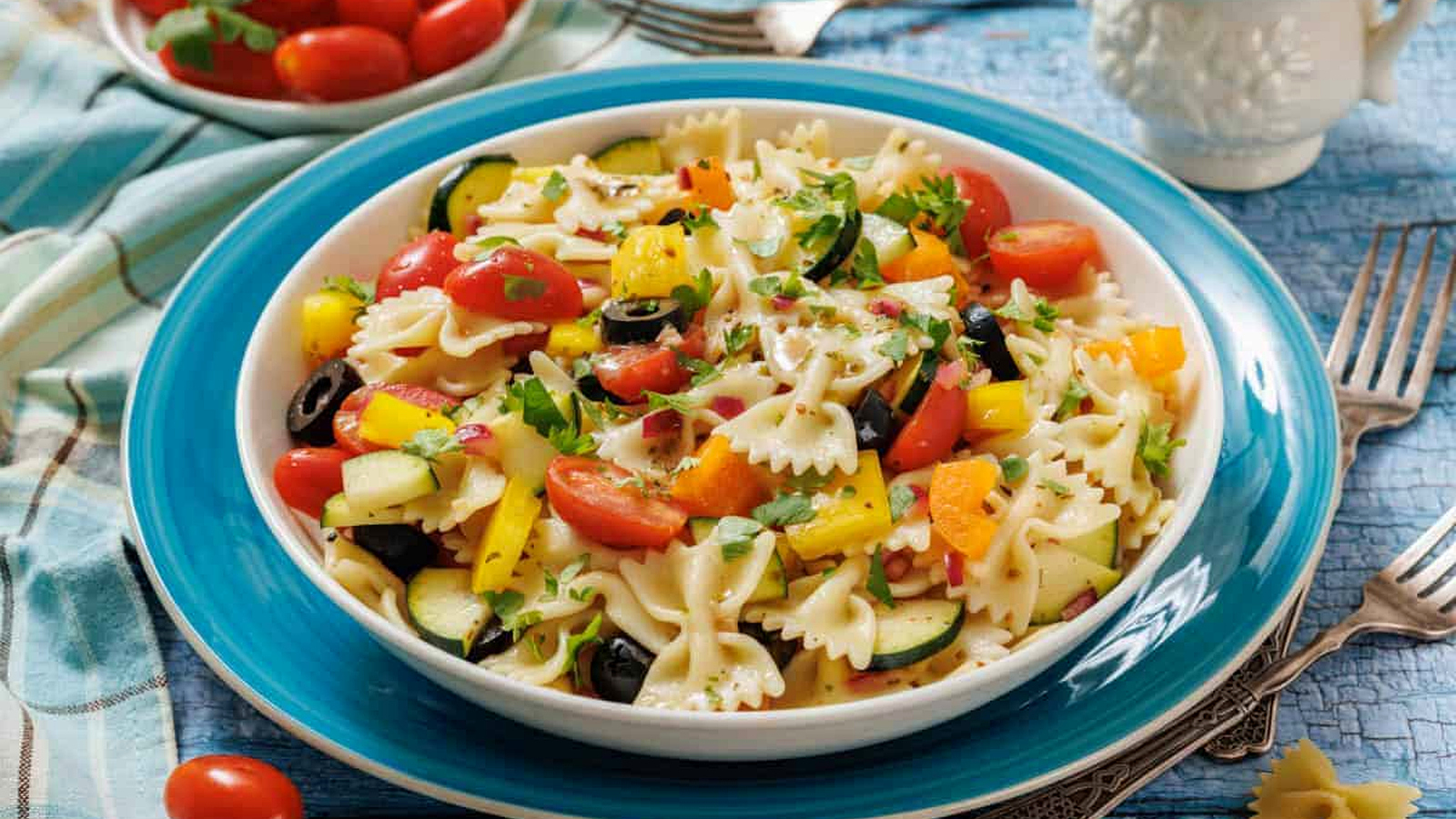 A plate of bowtie pasta salad with cherry tomatoes, yellow bell peppers, zucchini, black olives, and herbs, set on a blue plate with a bowl of tomatoes and utensils nearby.