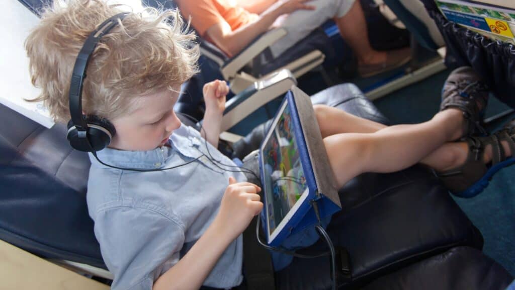 A young boy sits in an airplane seat wearing headphones, watching or playing on a tablet with his legs crossed.