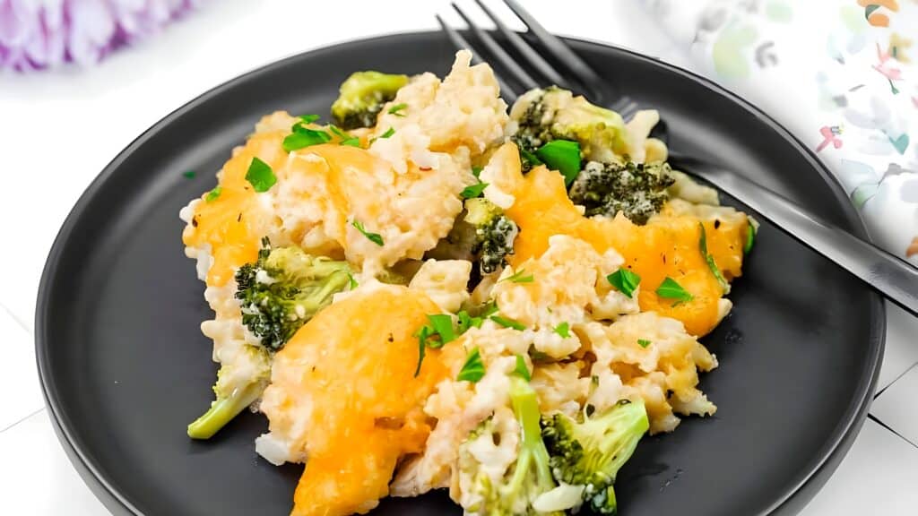 A black plate with a serving of cheesy broccoli and rice casserole, garnished with chopped parsley, with a fork placed on the side.