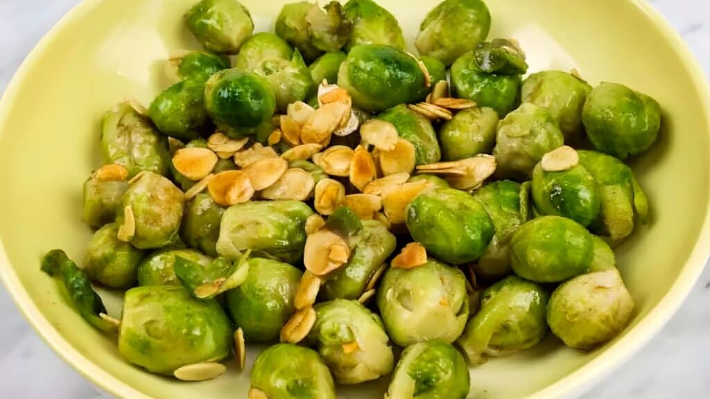 A bowl of cooked Brussels sprouts topped with sliced, toasted garlic on a light-colored surface.