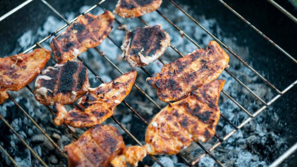 Several pieces of marinated meat are grilling on a metal grate over hot charcoal, showing perfect grill marks and slight charring&mdash;a true showcase of chef grilling tips in action.