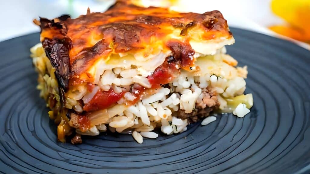 A close-up of a layered casserole slice on a dark plate, featuring browned cheese on top, with visible rice, ground meat, and tomato sauce layers.