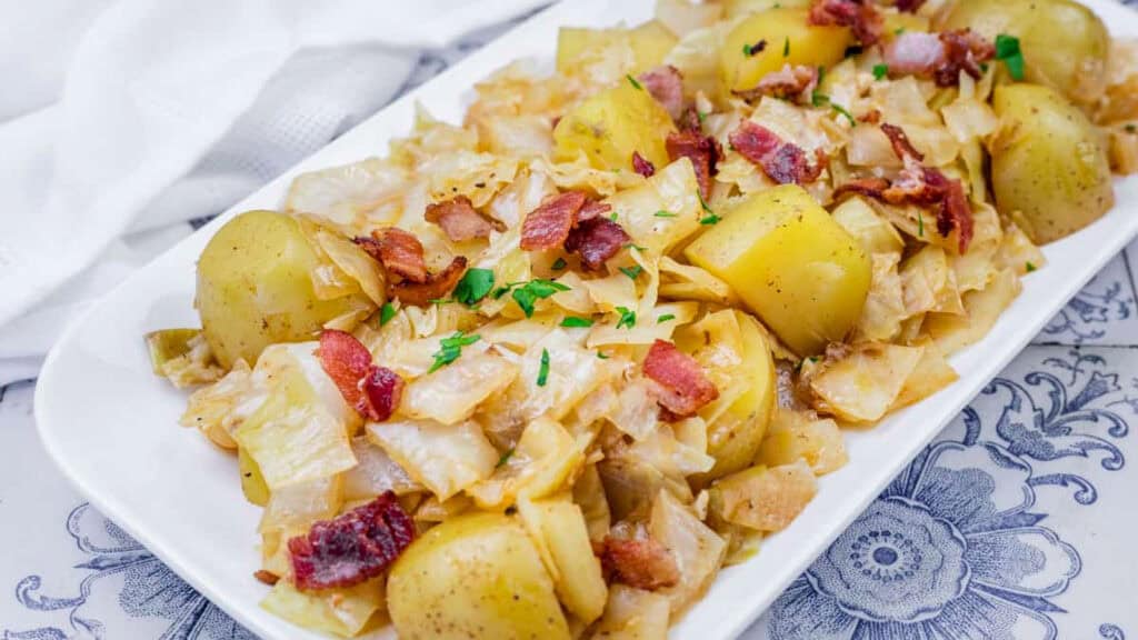 A white platter with cooked cabbage, potatoes, and bacon pieces, garnished with chopped parsley.