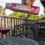 A hand pours red wine from a decorative can into a wine glass on an outdoor patio table with metal chairs.