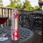 A glass of red wine and a bottle labeled "GAZE" sit on a metal patio table with chairs, on a wooden deck with trees and buildings in the background.