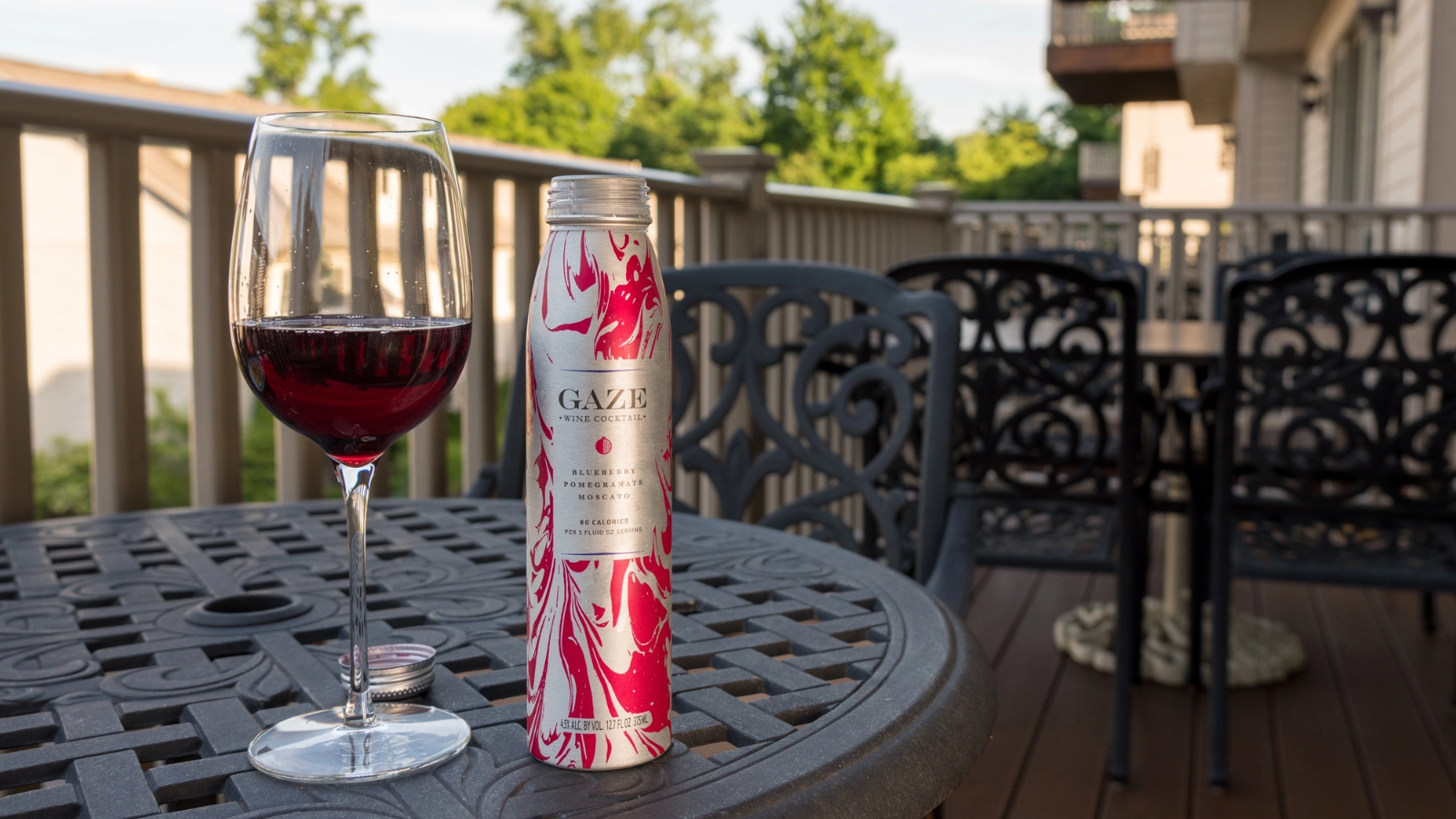 A glass of red wine and a bottle labeled "GAZE" sit on a metal patio table with chairs, on a wooden deck with trees and buildings in the background.