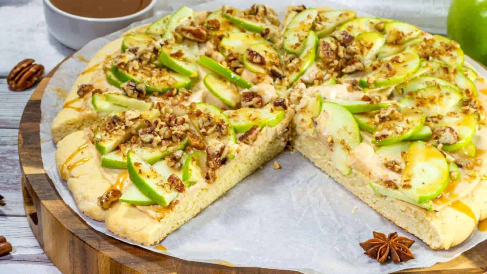 A dessert pizza topped with sliced green apples, chopped pecans, caramel drizzle, and cream cheese frosting, with one slice missing.