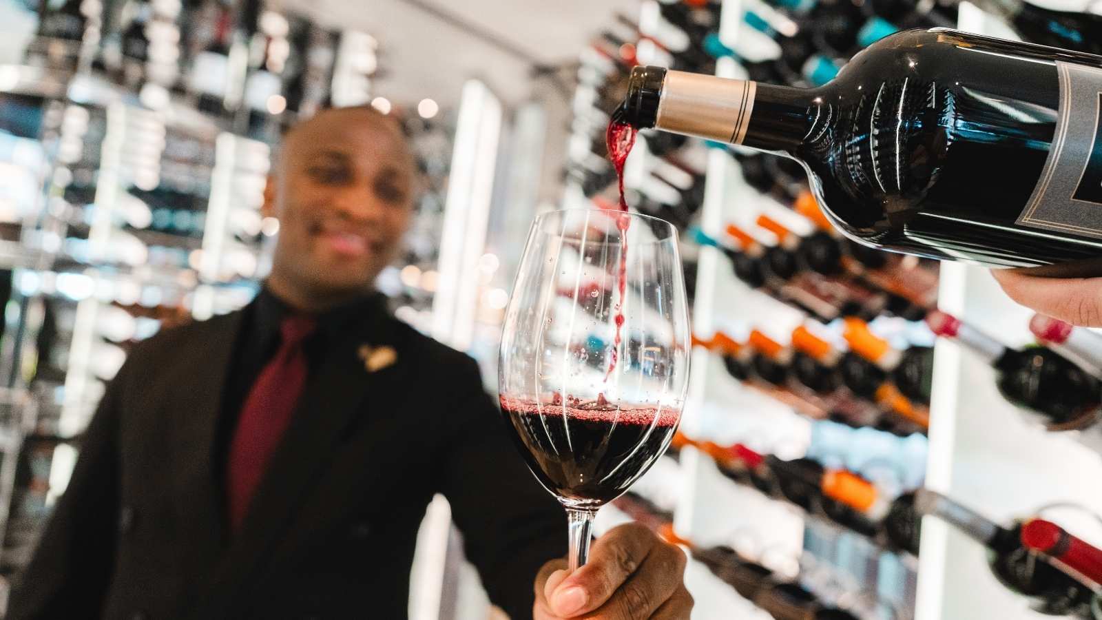 A person pours red wine into a glass held by a man in formal attire, with shelves of wine bottles in the background, reminiscent of a Celebrity Cruises wine tasting experience.