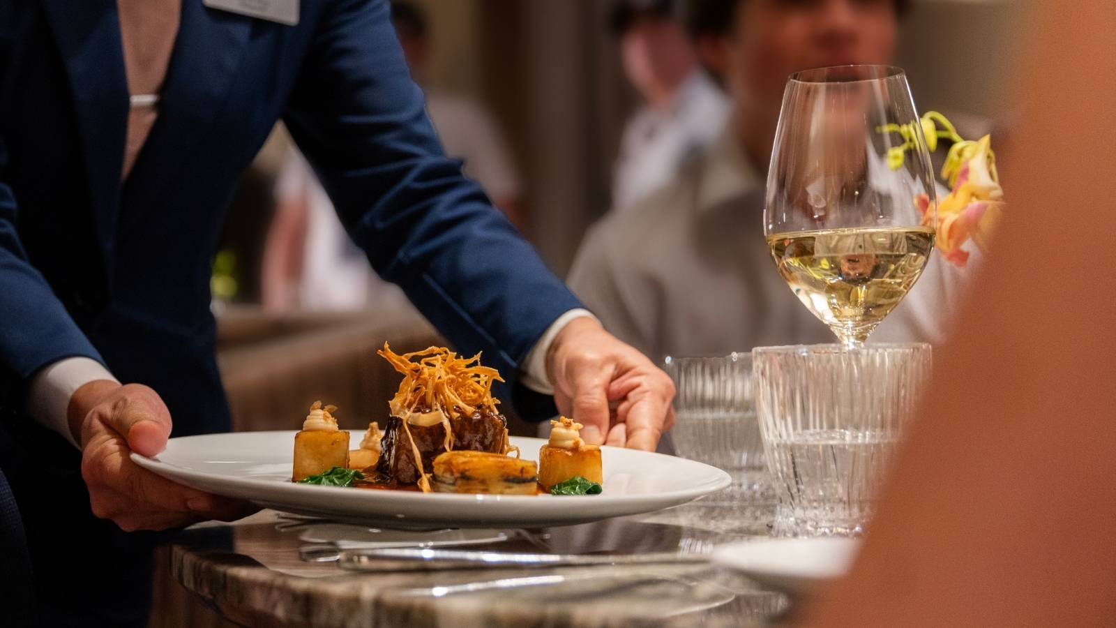 A server in a suit sets a plated gourmet dish on a marble table next to a glass of Celebrity Cruises wine in an elegant restaurant setting.
