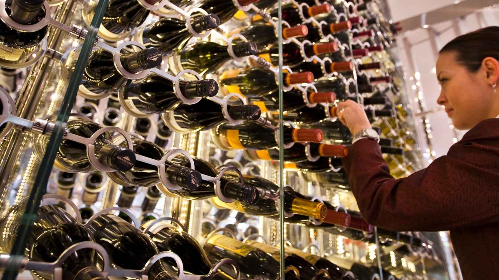 A person selects a wine bottle from a modern, glass-enclosed wine rack filled with horizontally stored bottles, reminiscent of the elegant selections found on Celebrity Cruises wine experiences.